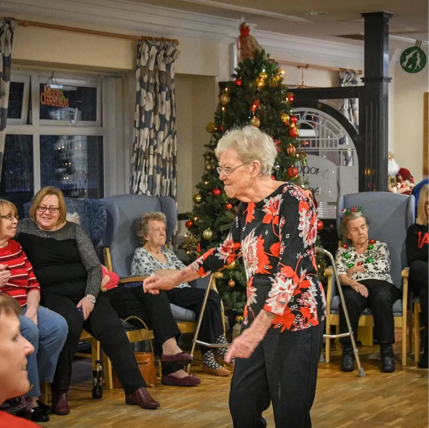 A resident dancing beside the Christmas tree in the lounge while others watch from their chairs.
